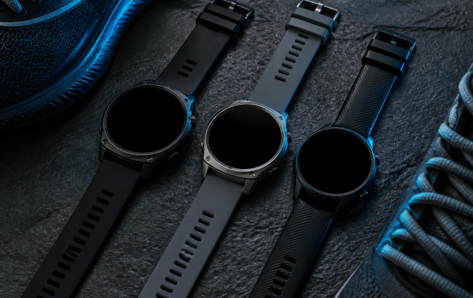 Drie ronde sport-smartwatches met zwarte bandjes op een donkere steen naast hardloopschoenen